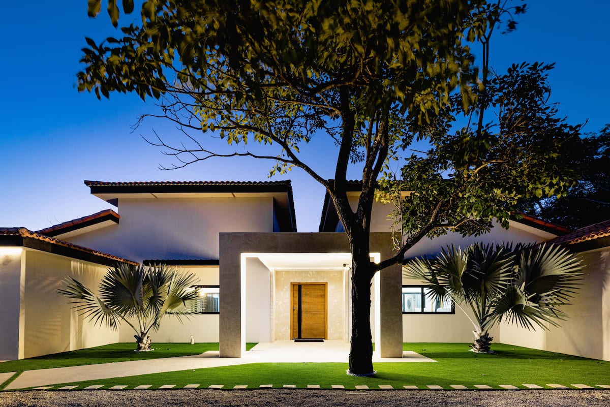 The entrance of Villa NAUA at dusk, under a tree, its front door lit
