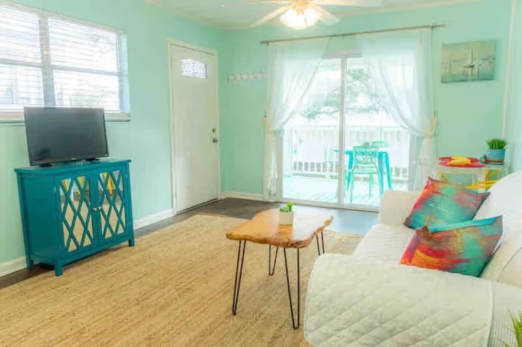 The living area features turquoise walls complemented by a light-colored sofa adorned with colorful accent pillows. A wooden coffee table is positioned centrally, with a smart TV on a blue cabinet nearby. Sheer curtains frame the glass door that leads to the covered porch.