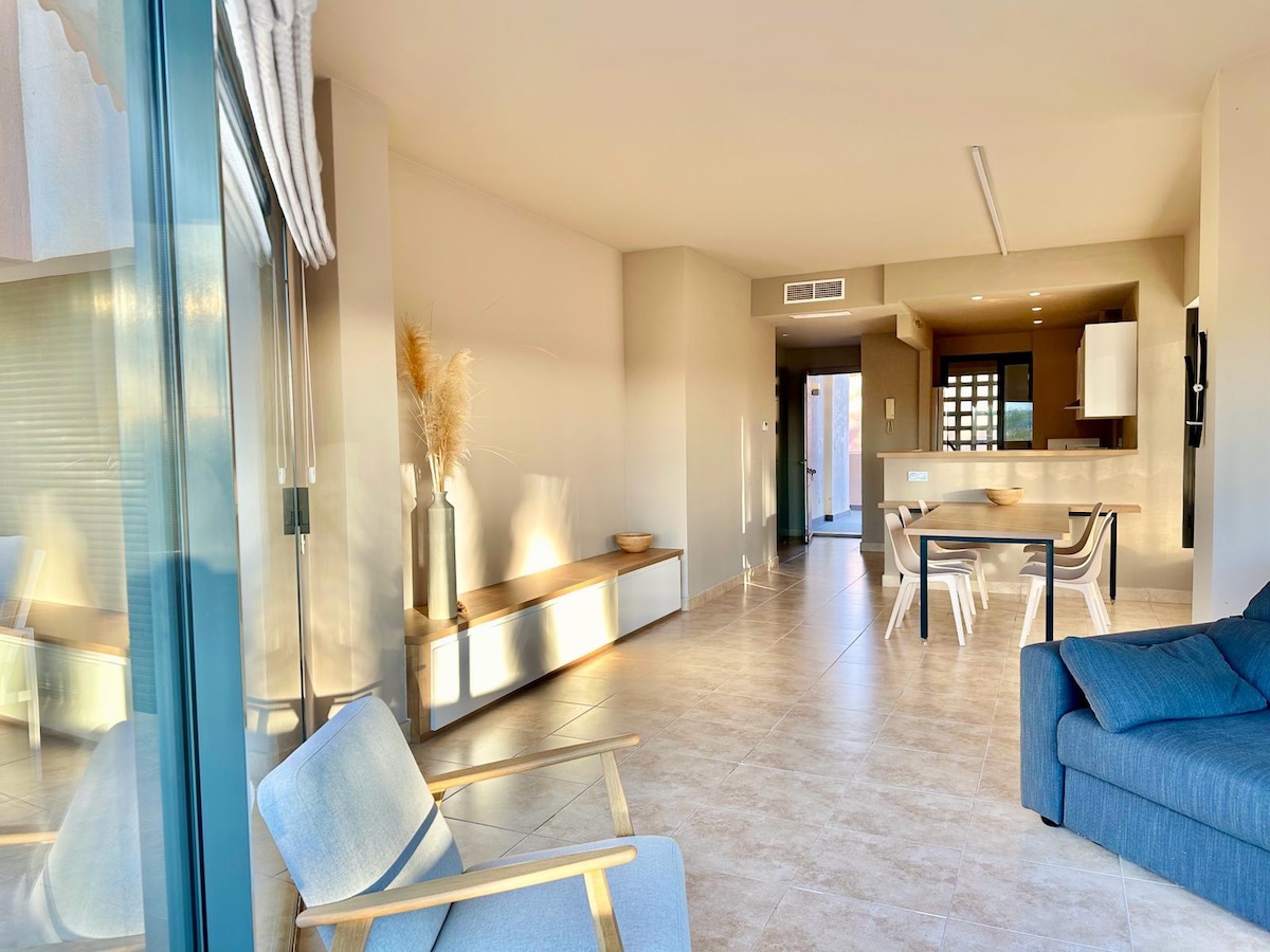 A spacious living area features large windows allowing ample natural light. A comfortable sofa sits alongside a minimalist dining table and chairs. The kitchen is visible in the background, equipped with modern appliances. A cozy corner with a decorative plant adds a touch of warmth.