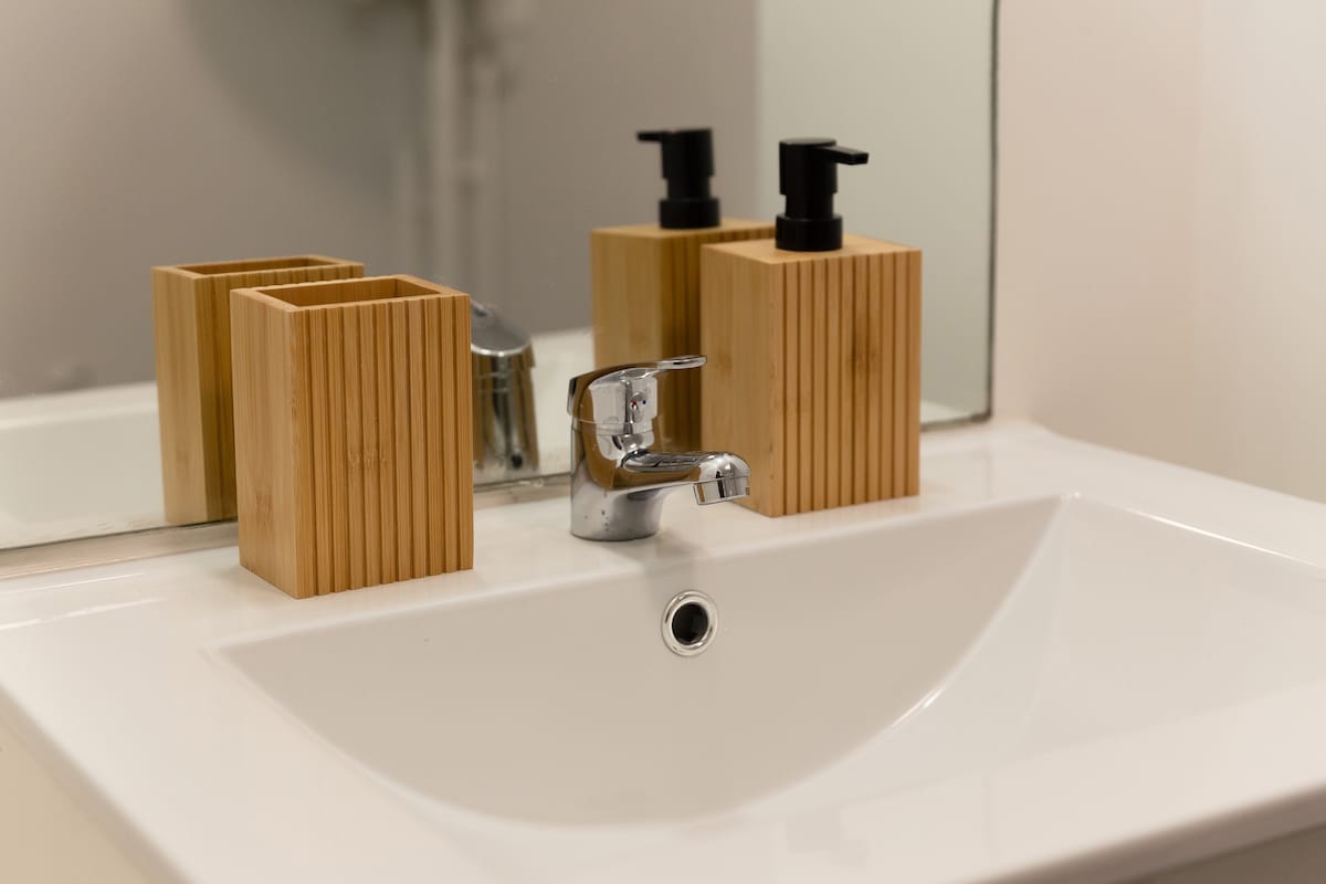 A modern bathroom sink is presented with a sleek chrome faucet and a clean white basin. Two wooden containers hold toiletries beside the faucet, offering a natural aesthetic. A mirror reflects the arrangement, enhancing the minimalist design and functionality of the space.