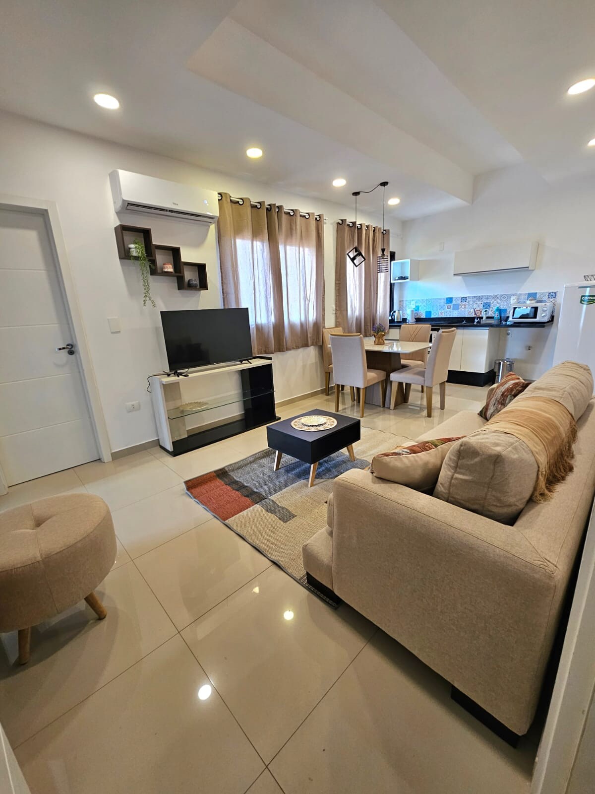 A light-filled living area features a comfortable sofa and a flat-screen TV. A dining table with four chairs is visible in the background, while a modern kitchen is equipped with essential appliances. Neutral tones are complemented by warm lighting and practical furniture arrangements.