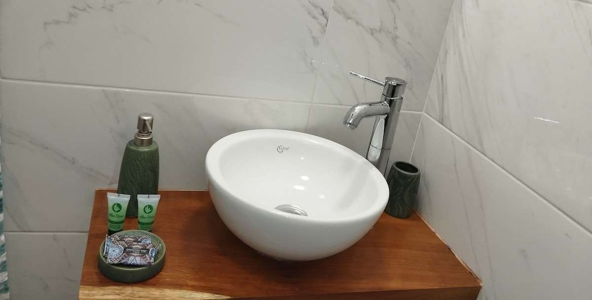 A modern bathroom features a sleek, round sink positioned on a wooden countertop. Decorative toiletries are arranged nearby, including a soap dish, lotion bottle, and small grooming products. The walls are adorned with marble tiles, enhancing the clean and contemporary look of the space.
