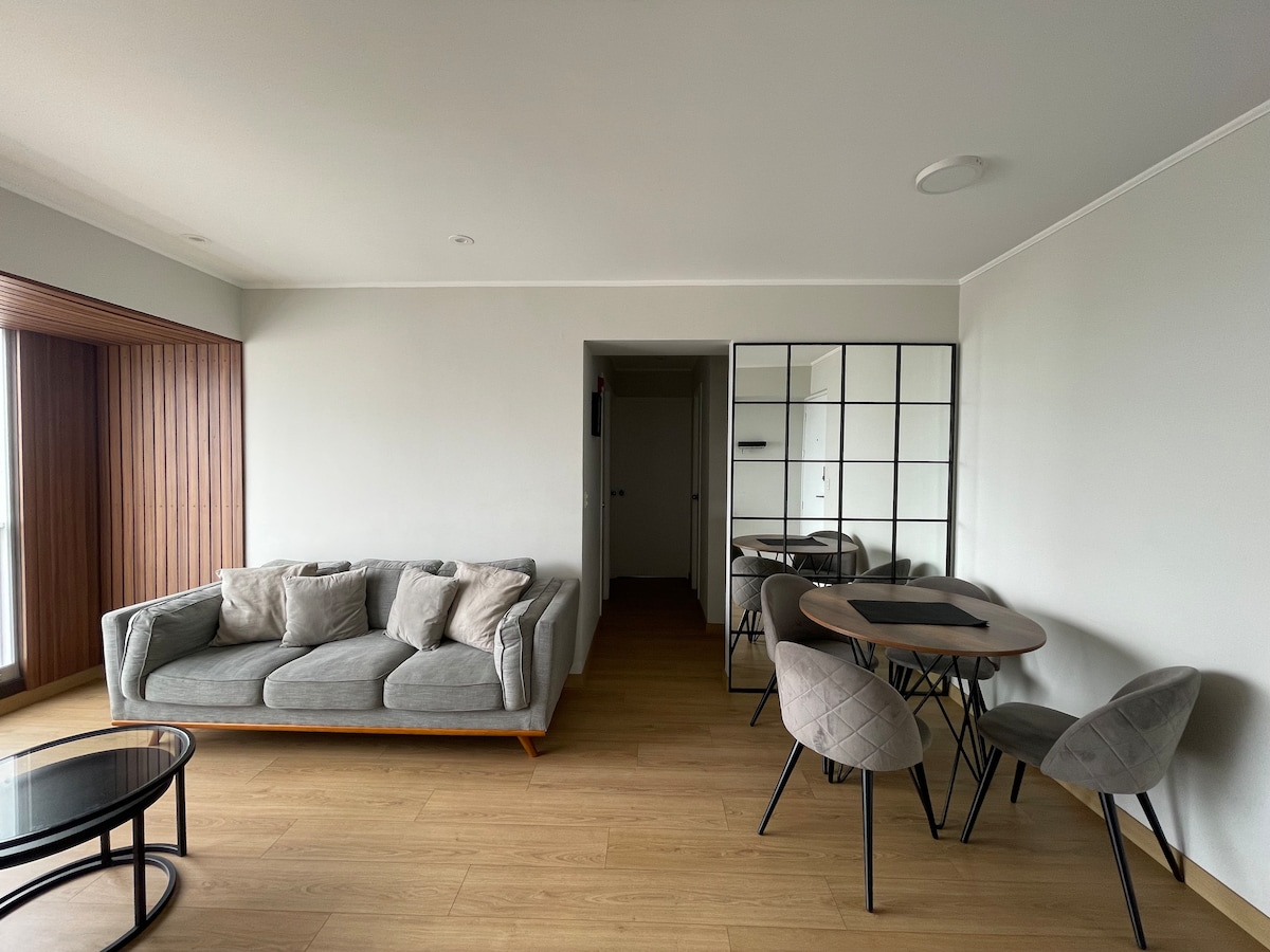 A spacious living area features a comfortable gray sofa adorned with cushions. A round dining table with four upholstered chairs is positioned nearby, complemented by a glass coffee table. A large mirror enhances the light in the room, and sliding glass doors lead to a scenic view.