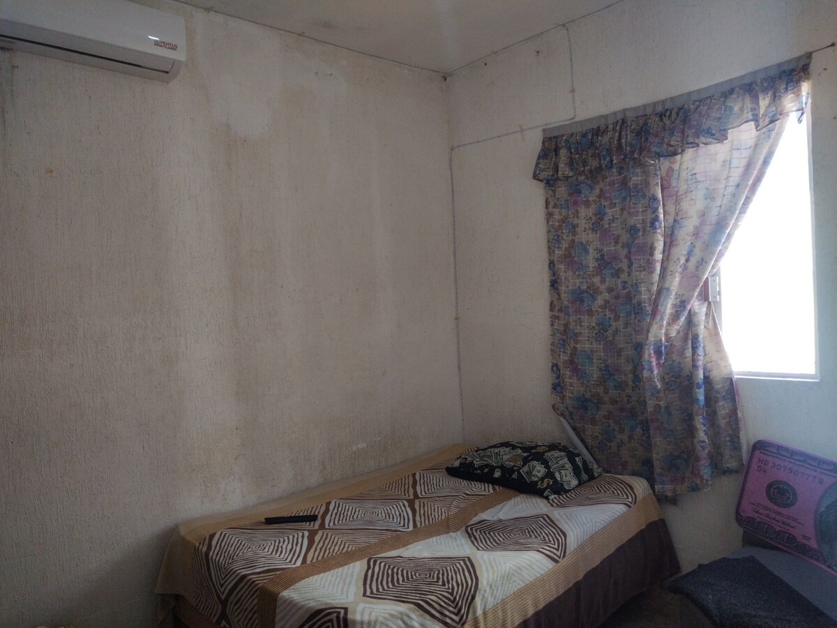 A simple bedroom features a single bed covered with a patterned blanket. A cushioned chair is positioned beside the bed. Floral curtains frame a window, allowing natural light to enter the space, while the walls exhibit a plain finish.