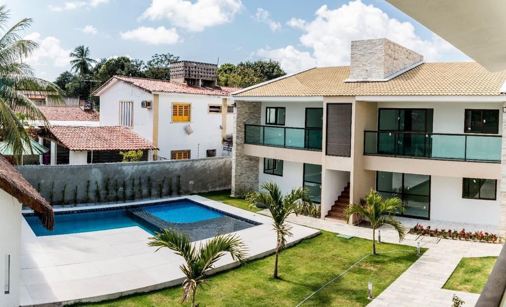 The exterior of the property features a modern two-story building with large glass balconies. A serene pool is situated in a well-maintained garden, bordered by palm trees and manicured grass, while neighboring houses can be seen in the background under a bright blue sky.