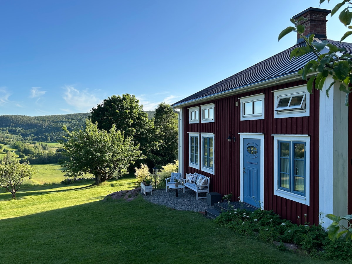 The exterior of a renovated guesthouse is presented, featuring deep red wooden siding and white accents. Multiple windows allow natural light to flood the interior. A welcoming blue door invites entry, framed by a small seating area. Lush greenery and hills are visible in the background.