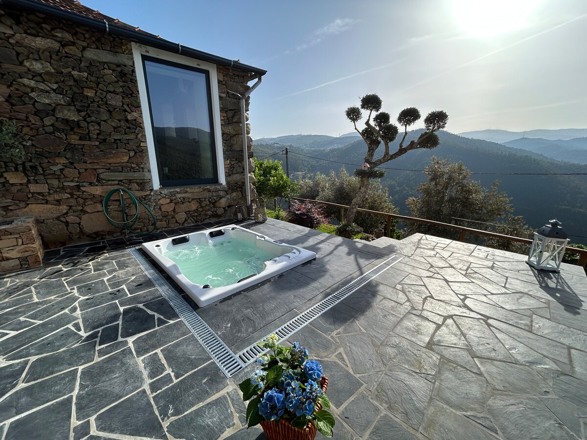 A private outdoor jacuzzi is positioned on a stone patio, surrounded by verdant hills and trees. The sun illuminates the landscape, while a flower arrangement adds a touch of color to the scene, inviting relaxation amidst nature's tranquility.