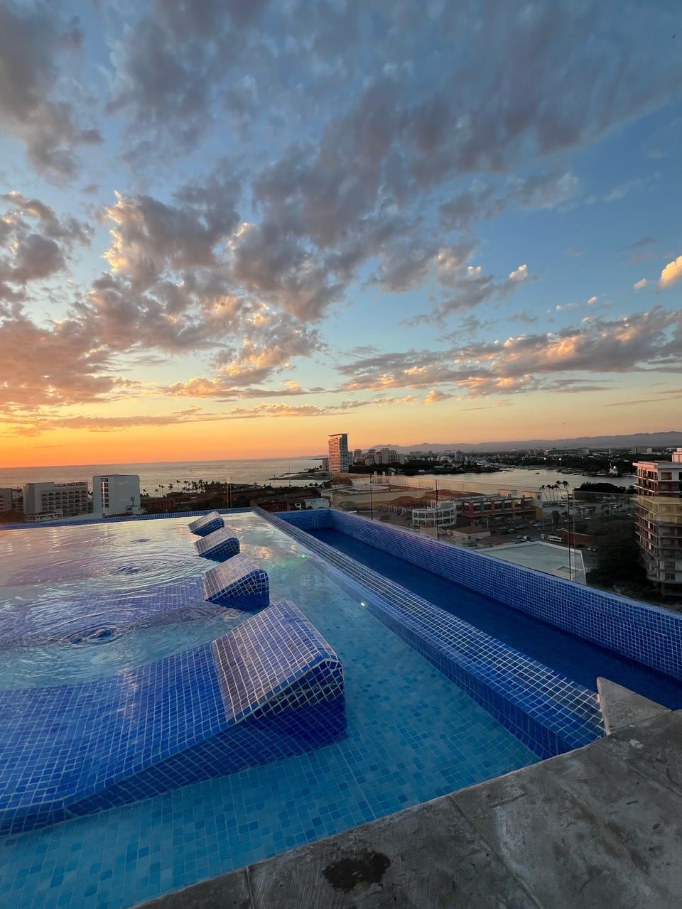 An infinity pool is presented against a backdrop of a colorful sunset sky. Soft waves ripple across the water's surface, reflecting shades of orange, pink, and blue. The surrounding cityscape and coastline are visible in the distance, contributing to a serene atmosphere.