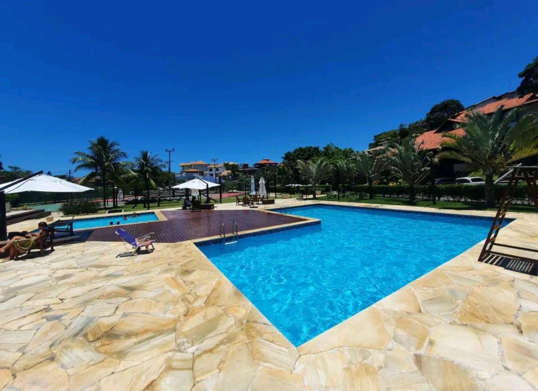 A spacious outdoor area features two swimming pools surrounded by lush palm trees. Lounge chairs are positioned under umbrellas on the stone patio. Clear blue skies provide a bright backdrop for the inviting pools, enhancing the atmosphere of relaxation.