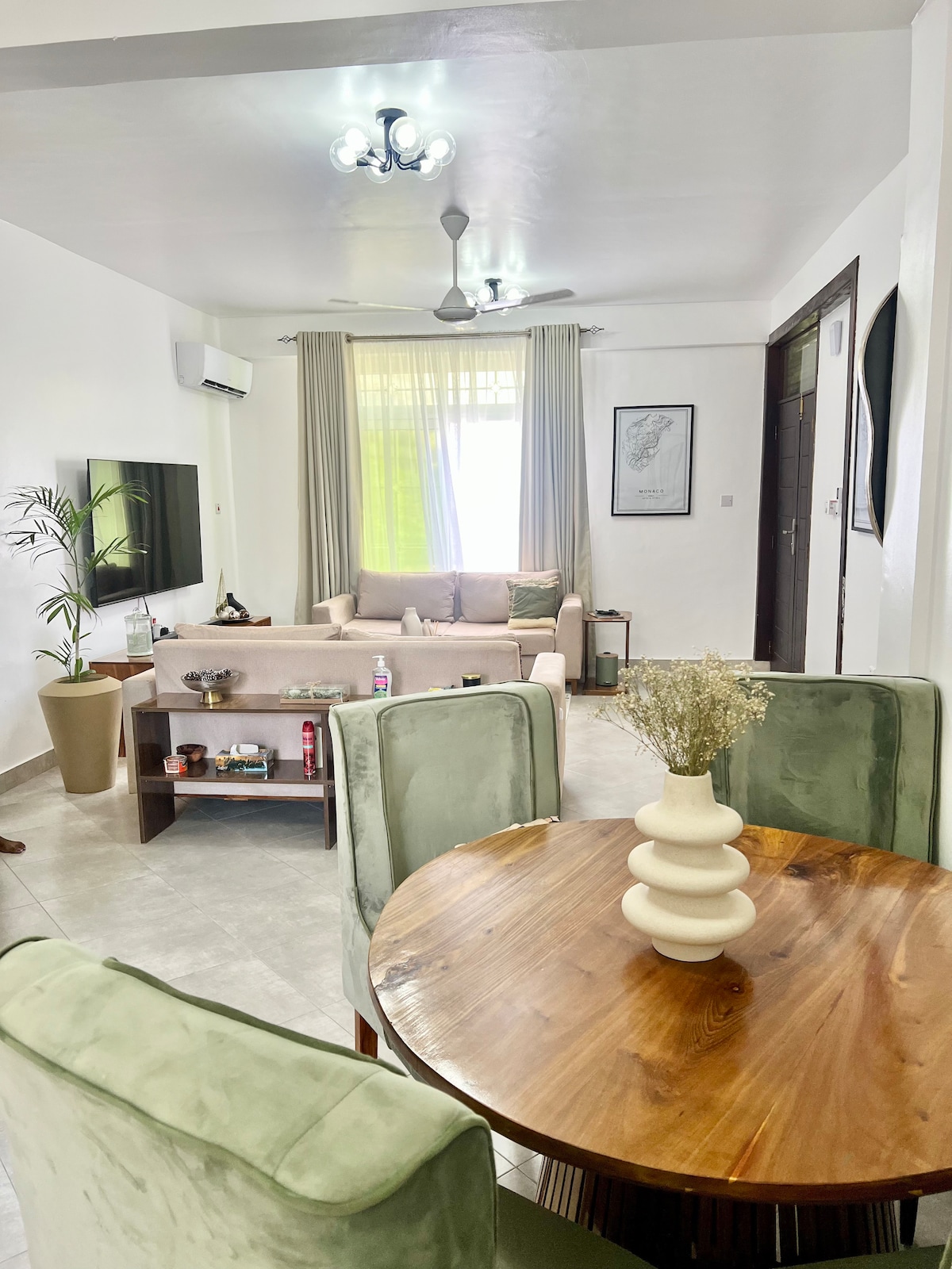 A modern living area features a comfortable seating arrangement with a light-colored sofa and decorative wall art. A round wooden table surrounded by soft green chairs offers an inviting dining space. Large windows allow ample natural light, complemented by stylish light fixtures.