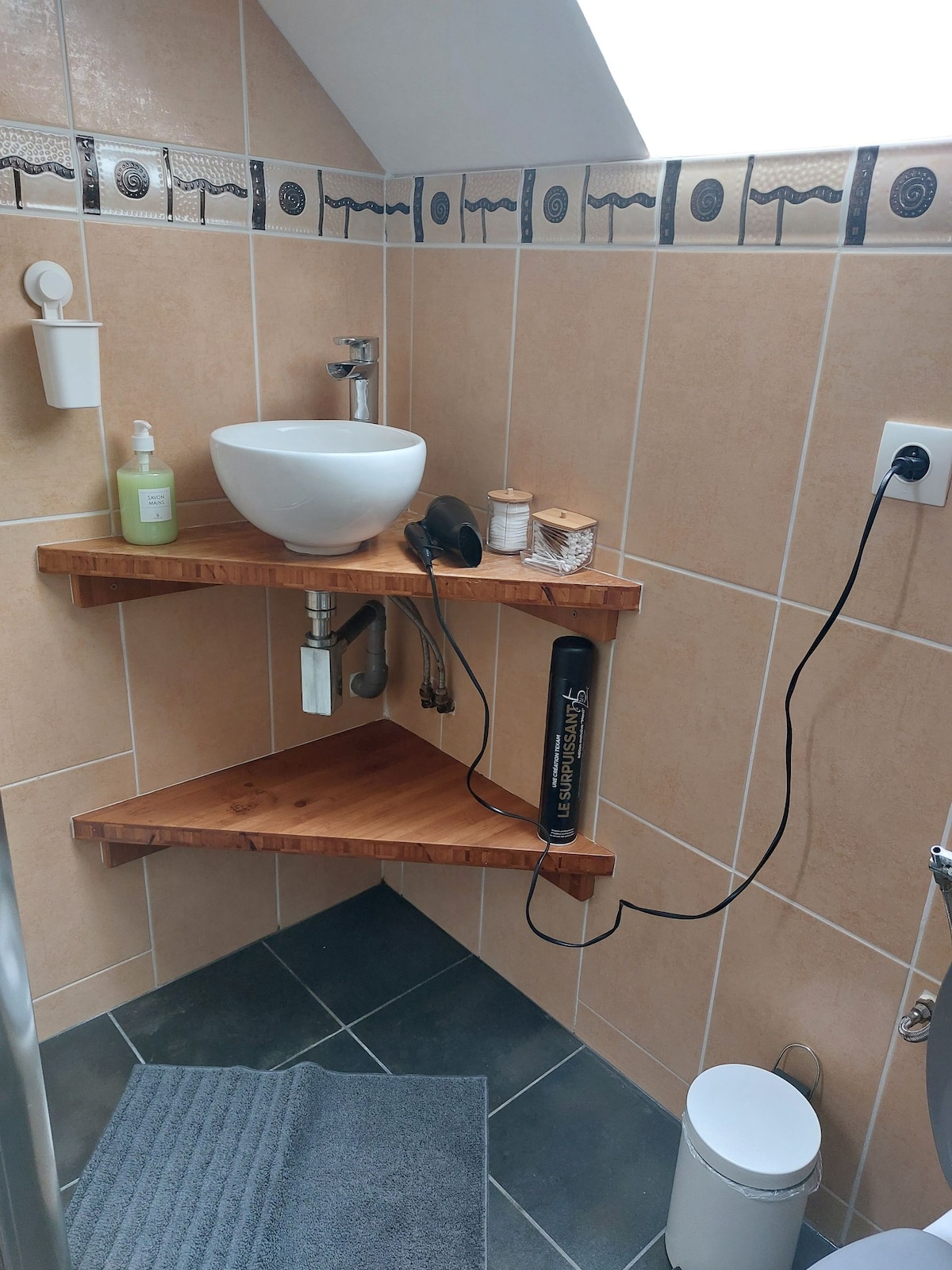 A modern bathroom area features warm wooden shelves that hold toiletries and a basin, complementing the light-colored tiled walls. A gray bath mat is positioned near the sleek shower, while a small waste bin is tucked neatly in the corner.