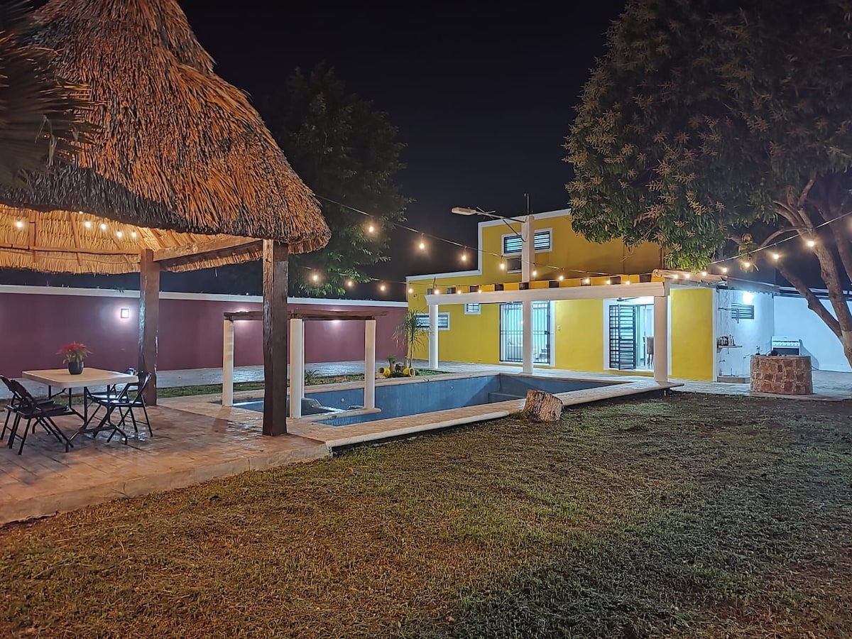 The outdoor area features a swimming pool bordered by a stone deck, illuminated softly by string lights overhead. A thatched-roof palapa provides shade for outdoor seating, while a vibrant yellow house stands in the background, showcasing large glass doors that lead outside.