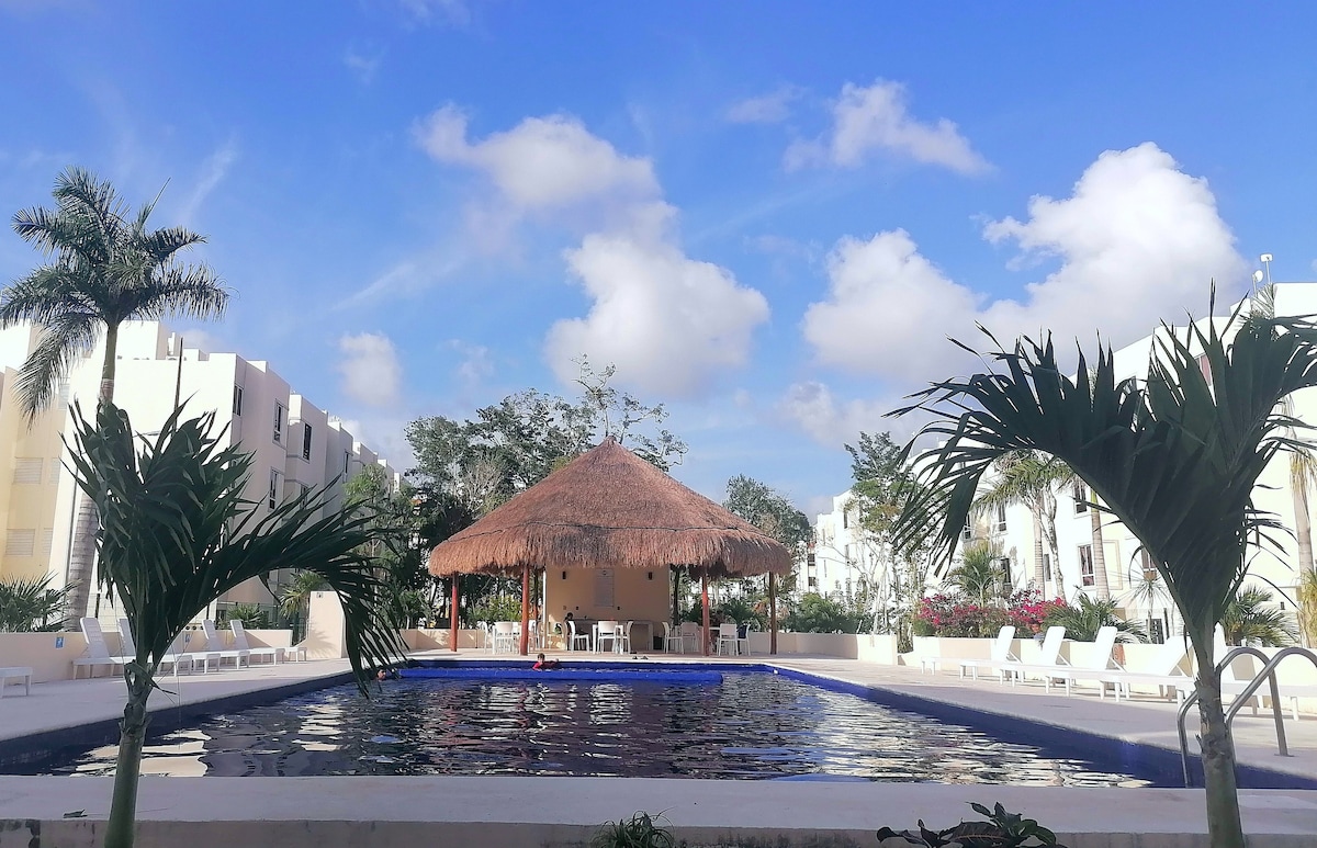 A shared swimming pool is surrounded by tropical greenery and features a thatched-roof gazebo at the center. Sun loungers are positioned along the poolside, under a bright blue sky dotted with fluffy white clouds.