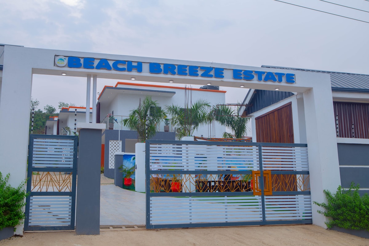 An entrance gate with the text 'Beach Breeze Estate' prominently displayed is featured. A landscaped pathway leads into the property, flanked by greenery and palm trees. The modern design of the estate is visible in the background, complementing the welcoming atmosphere.