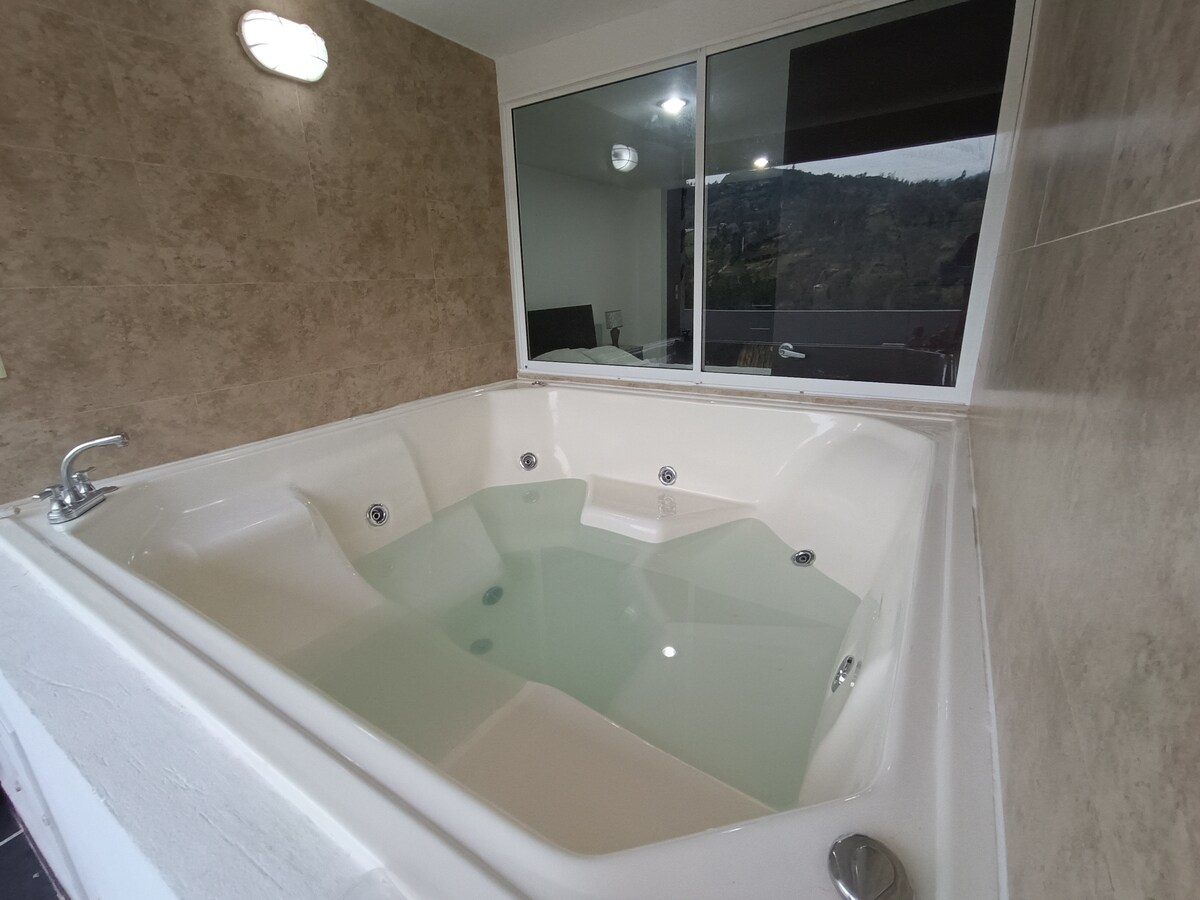 A spacious jacuzzi is situated in a well-lit corner, surrounded by textured beige walls. The inviting water surface reflects light from overhead fixtures, while a large window offers a glimpse of the outdoor view.