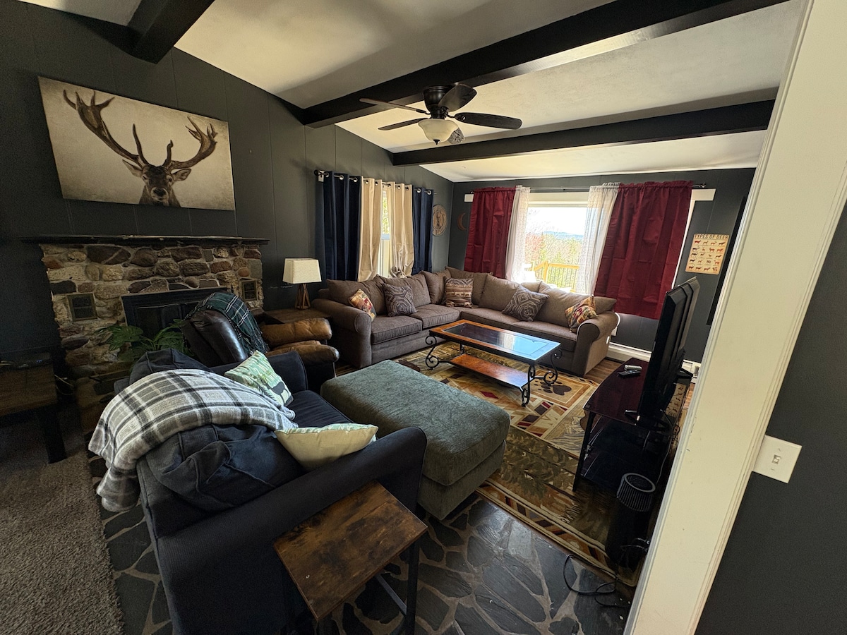 A spacious living room features a large sectional sofa with assorted cushions, a dark wood coffee table, and a stone fireplace adorned with an antler decoration. Natural light fills the room through a window framed by red and neutral drapes, enhancing the cozy atmosphere.