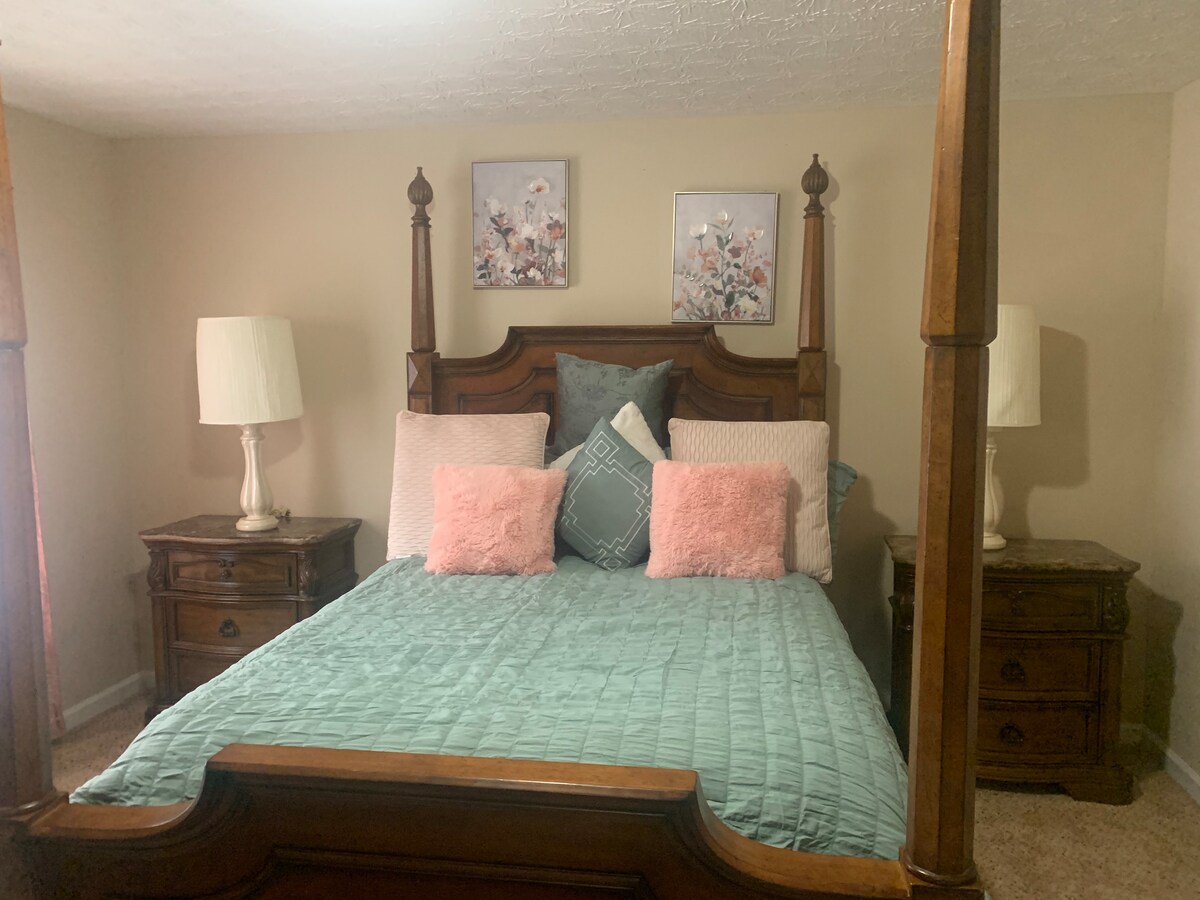 A spacious bedroom features a four-poster bed with a light blue quilt and decorative pillows in soft shades. Two bedside tables with lamps flank the bed, complemented by floral artwork on the walls, enhancing the calming atmosphere.