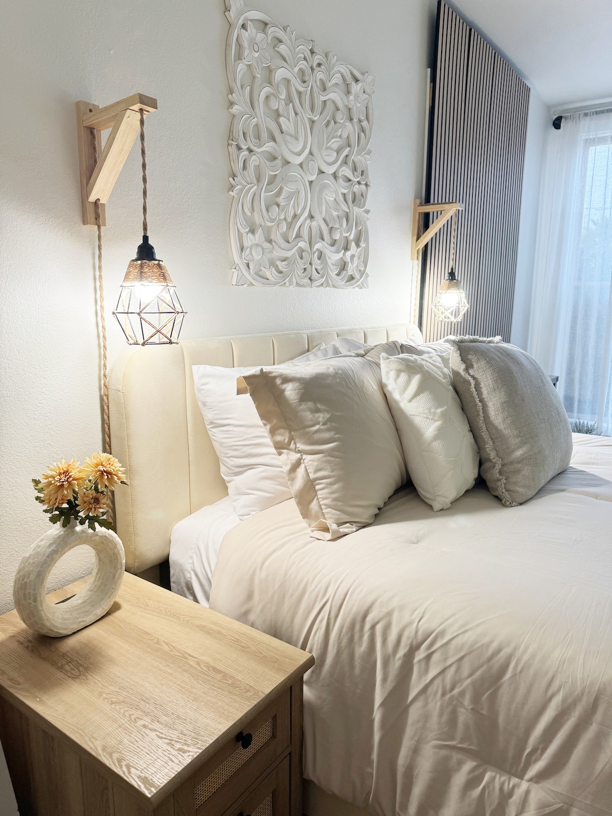 A well-appointed sleeping area is presented with a spacious bed featuring soft white linens and decorative pillows. Flanking the bed are bedside tables with wall-mounted lights, and a piece of wall art adds a touch of style. Natural light filters through sheer curtains.
