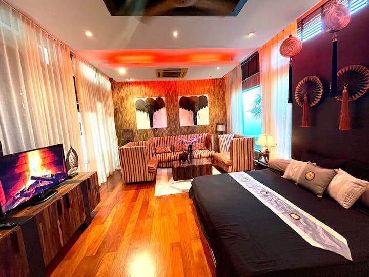Amplia recámara/sala estilo tailandés. En primer plano hay una cama grande con sábanas oscuras. En la zona de estar hay sofás a rayas, una mesa de madera y un televisor. Hay cabezas de elefante en las paredes y decoración asiática. Iluminación cálida y piso de madera.