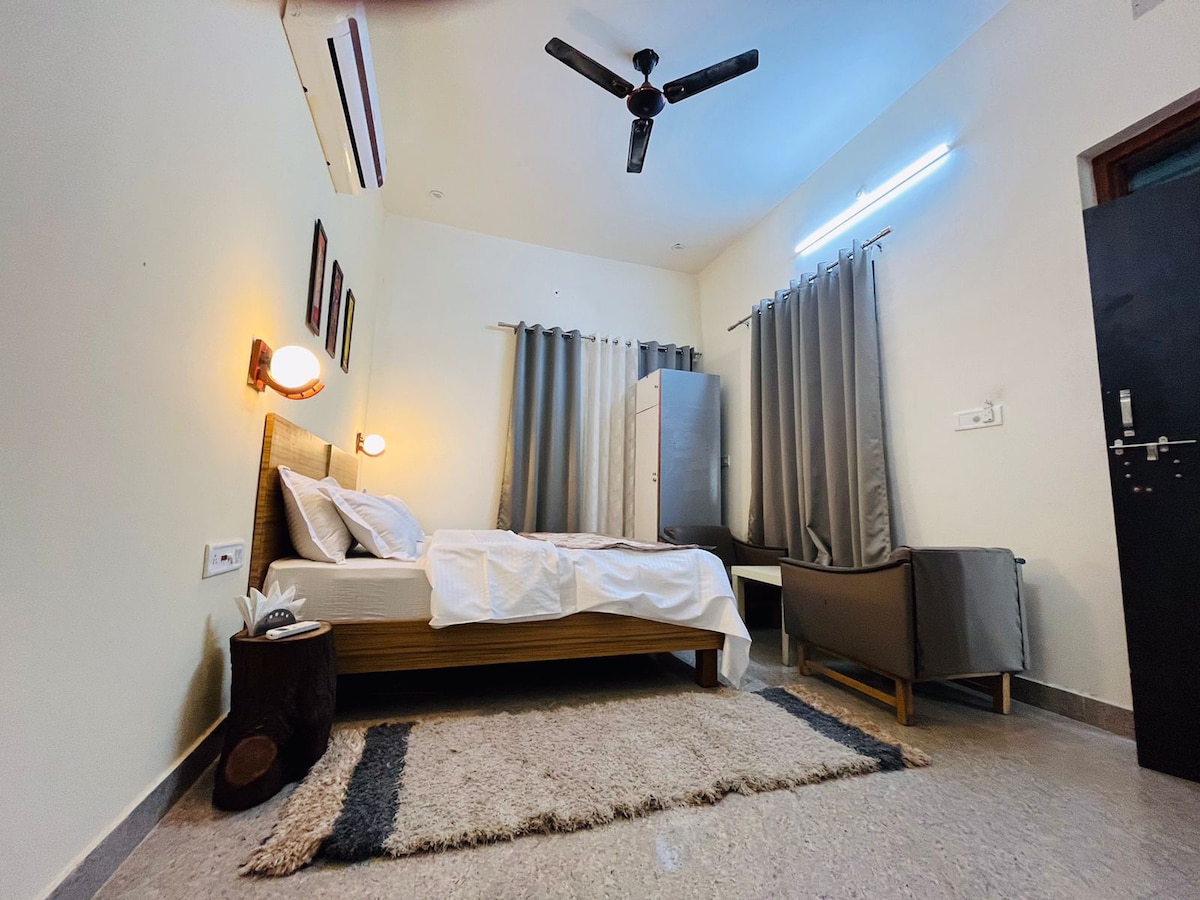 The room features a spacious bed with crisp white linens, complemented by a dark wood headboard. A gray sofa sits in one corner, and gray curtains frame a window. A light rug covers the floor, while wall-mounted lamps provide soft lighting.