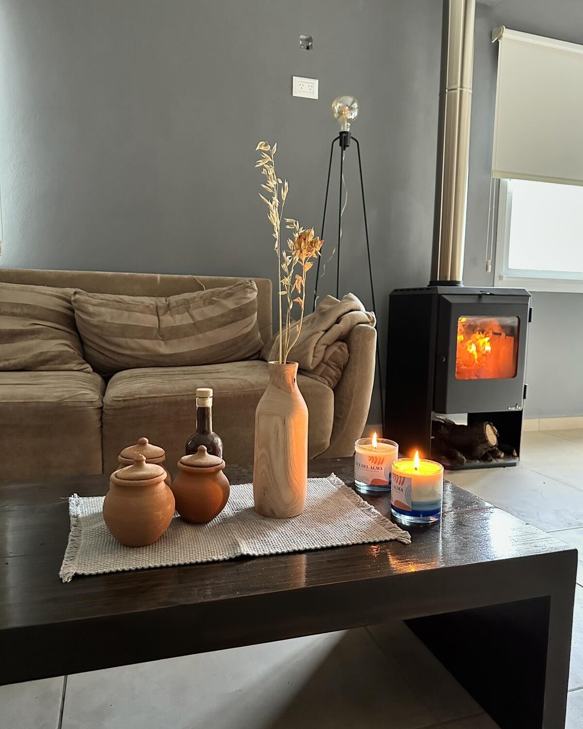 A comfortable living area features a neutral-colored couch next to a coffee table adorned with decorative items. Candles and a wooden vase with dried flowers are placed on a textured runner. A small fireplace provides warmth and ambiance, enhancing the cozy setting.