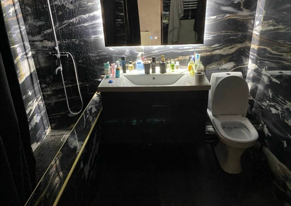 A modern bathroom features sleek black and gold marble walls. A vanity with a wide mirror and soft lighting is complemented by a variety of toiletries. A shower area is visible, with a glass panel and showerhead, alongside a standard white toilet.