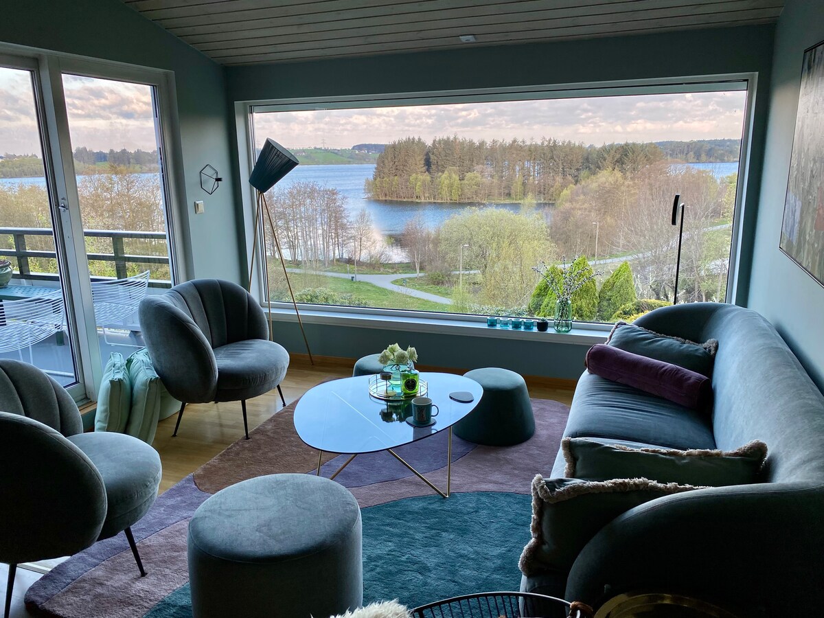 A bright living space features a large window showcasing a scenic view of Stokkavannet. Comfortable lounge chairs and a modern sofa surround a glass coffee table. Soft area rugs add warmth, while decorative accents enhance the inviting environment.