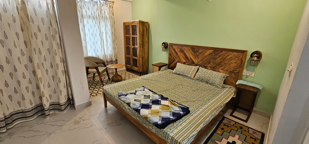 A spacious bedroom features a large wooden bed adorned with patterned bedding. Two bedside tables with lamps sit beside the bed. Natural light enters through a window, partially covered by sheer curtains. A wooden wardrobe and a cozy seating area are visible in the background.