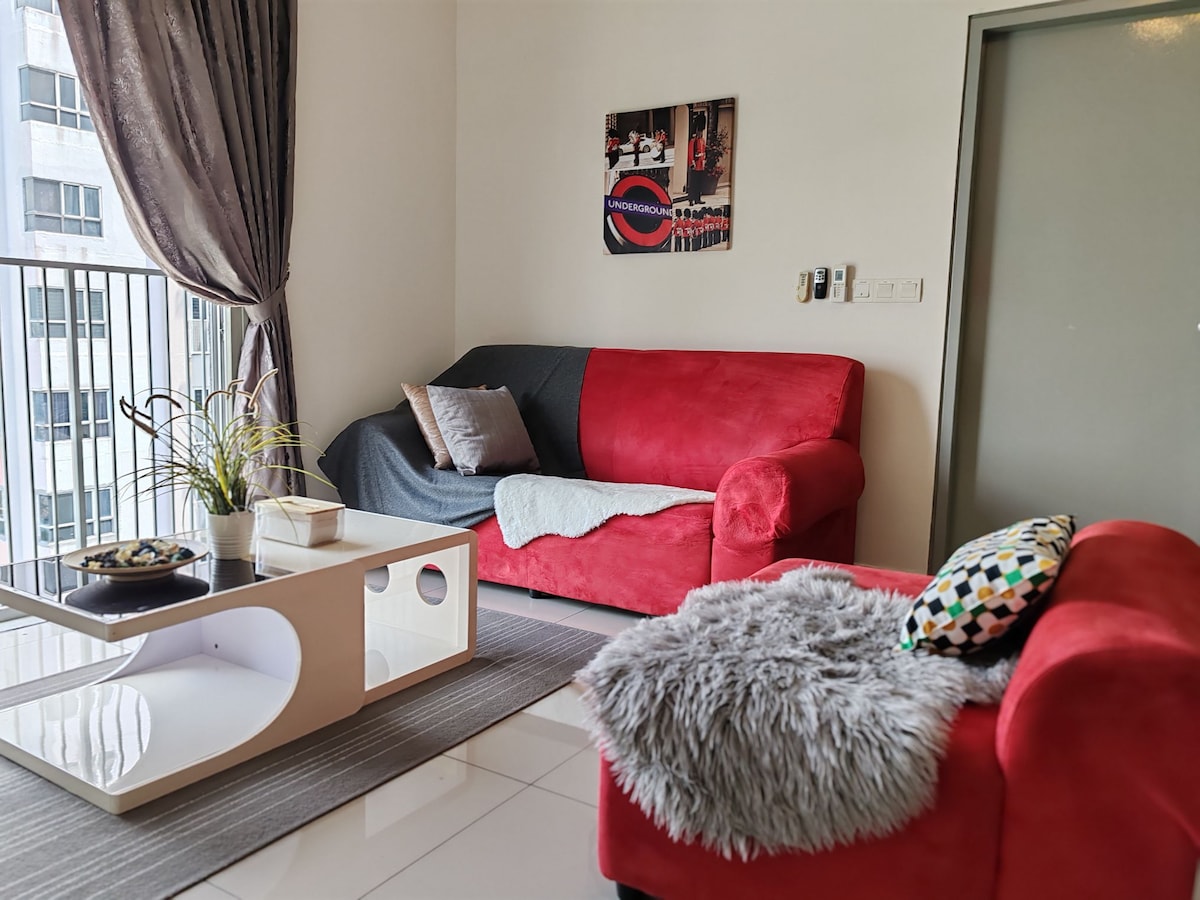 A cozy living area features a red L-shaped sofa adorned with a grey throw and a colorful patterned cushion. A modern coffee table with a smooth surface holds a decorative bowl. Natural light floods the space through a glass balcony door, complemented by floor-to-ceiling curtains.