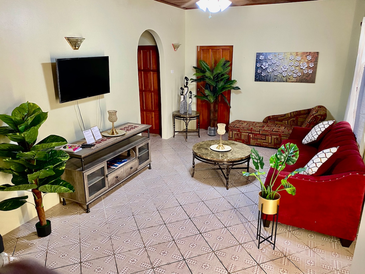 A welcoming living area features a television mounted on the wall, alongside a console table with decorative accents. A red sofa and patterned chair provide seating, complemented by a round coffee table. Green plants add a touch of nature, contributing to the overall comfort of the space.