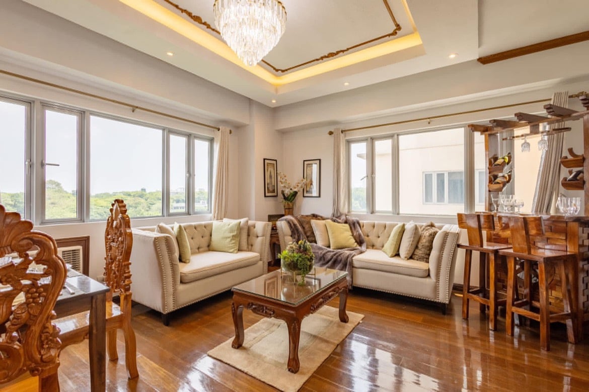 A bright living area features large windows that provide natural light and views of the outside. Comfortable sofas are arranged around a wooden coffee table, while a dining space with elegant chairs is visible in the background. A chandelier hangs from the ceiling, adding a touch of sophistication.