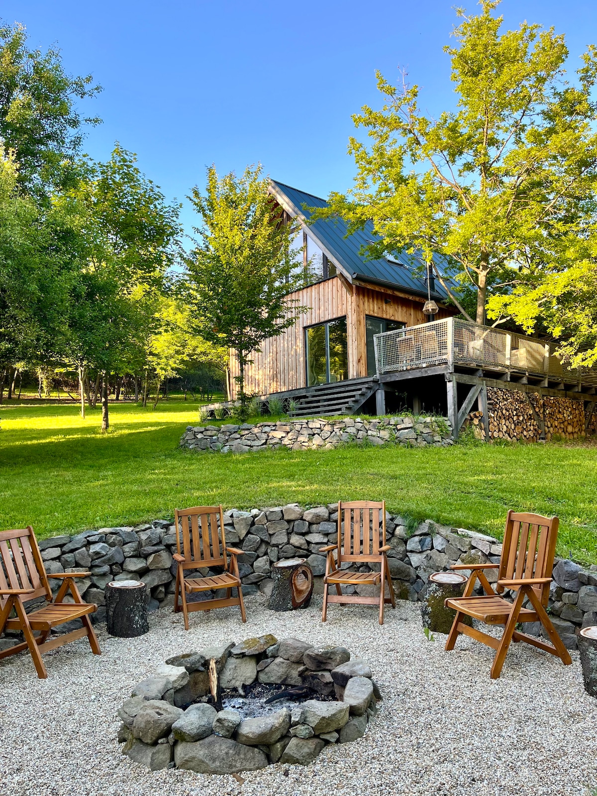 An outdoor seating area is arranged around a stone fire pit, featuring wooden chairs set on a gravel surface. A wooden house with a sloping roof and large windows is positioned in the background, surrounded by lush green trees and an expansive lawn.