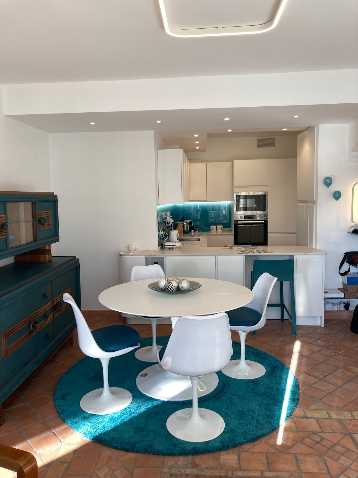 A modern dining area is highlighted by a round table surrounded by four ergonomic chairs. A vibrant blue rug adds a pop of color to the terracotta tiled floor. The kitchenette, featuring sleek cabinetry and appliances, is visible in the background.