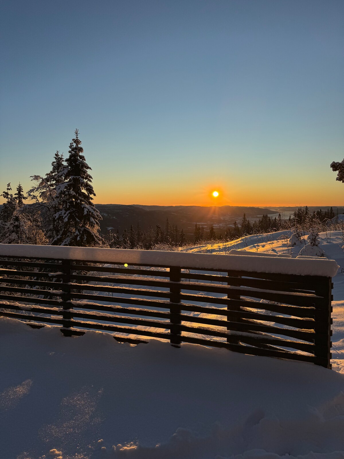A snow-covered landscape is bathed in the warm light of a sunset, with a wooden railing framing the view. Snow-laden trees can be seen in the foreground, while rolling hills stretch into the distance under a golden sky.