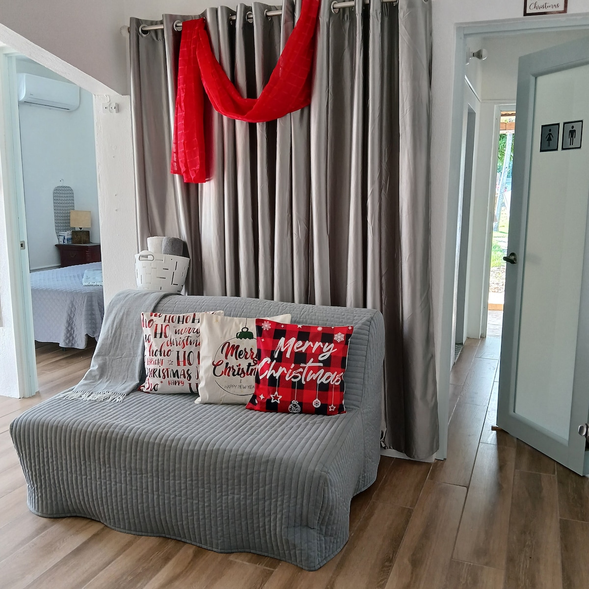 A cozy living area features a soft gray sofa adorned with decorative pillows, including a festive 'Merry Christmas' design. Light wooden flooring complements the space, where natural light is allowed to flow through a nearby doorway leading to other parts of the home.
