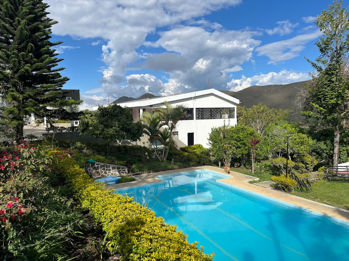 A serene view of the pool is complemented by lush gardens and blooming flowers. The house features large windows that invite natural light, with distant mountains framing the landscape under a vibrant sky. Sun loungers are positioned around the pool.