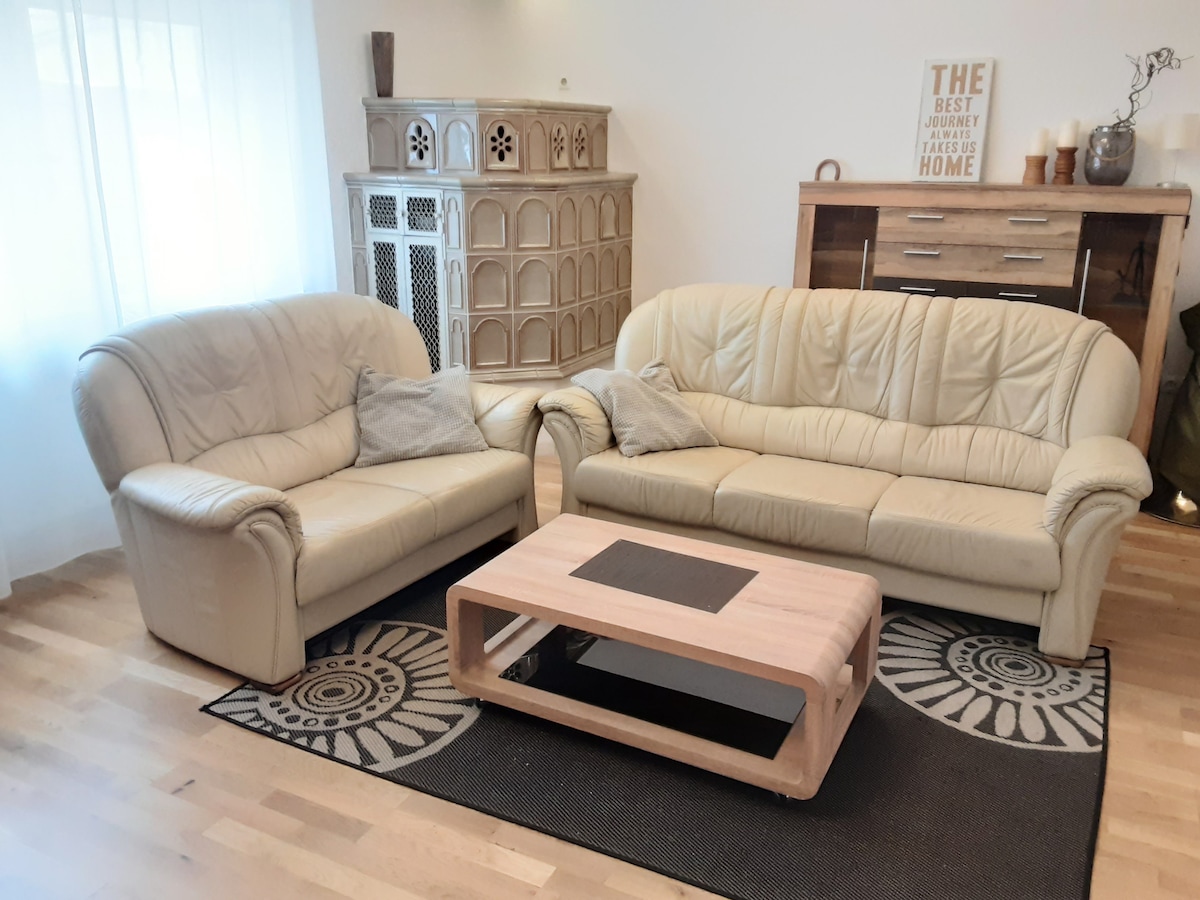 A light-filled living area features two cream leather sofas positioned around a wooden coffee table. A decorative area rug with a geometric pattern is placed underneath, while neutral walls and well-placed decor add a harmonious touch to the space.