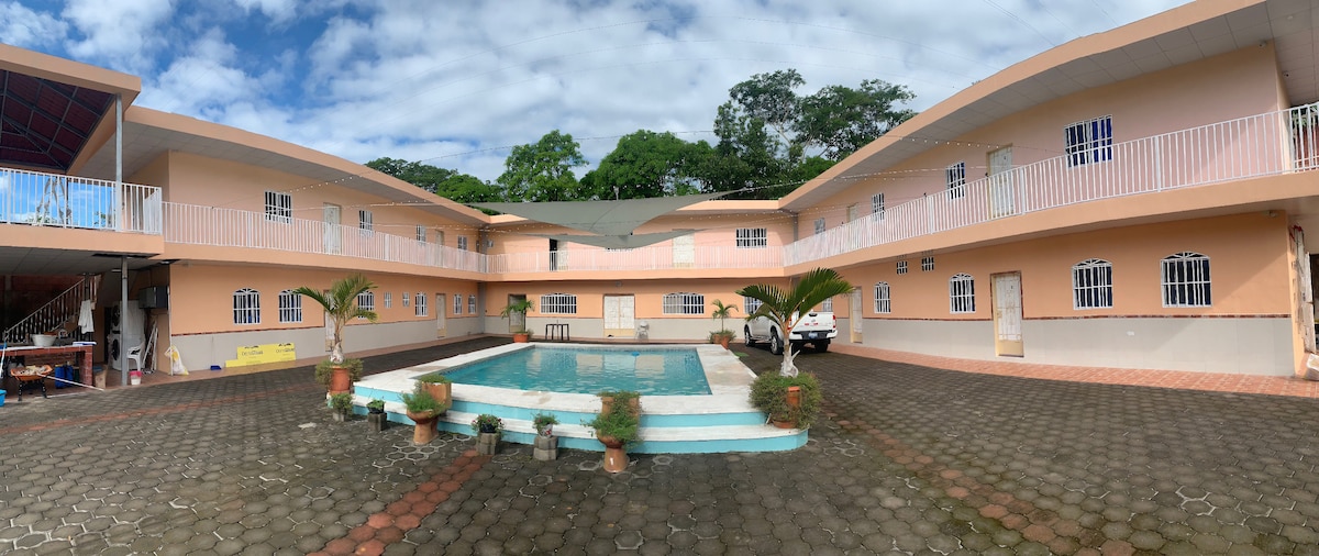 An outdoor pool surrounded by a paved courtyard is framed by buildings with a light peach facade. Lush greenery and potted plants enhance the tranquil setting. Balconies with railing are visible on the upper level, providing additional access to the inviting outdoor space.