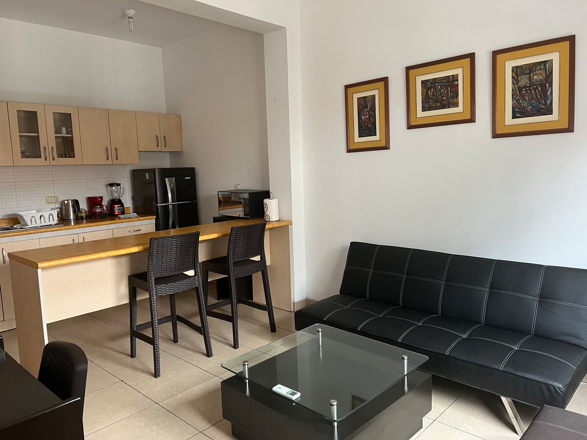 An open-plan living area features a sleek black sofa, a glass coffee table, and a dining space with two bar stools. The kitchen area is equipped with modern appliances, and decorative artwork is displayed on the walls, contributing to a functional and stylish ambiance.