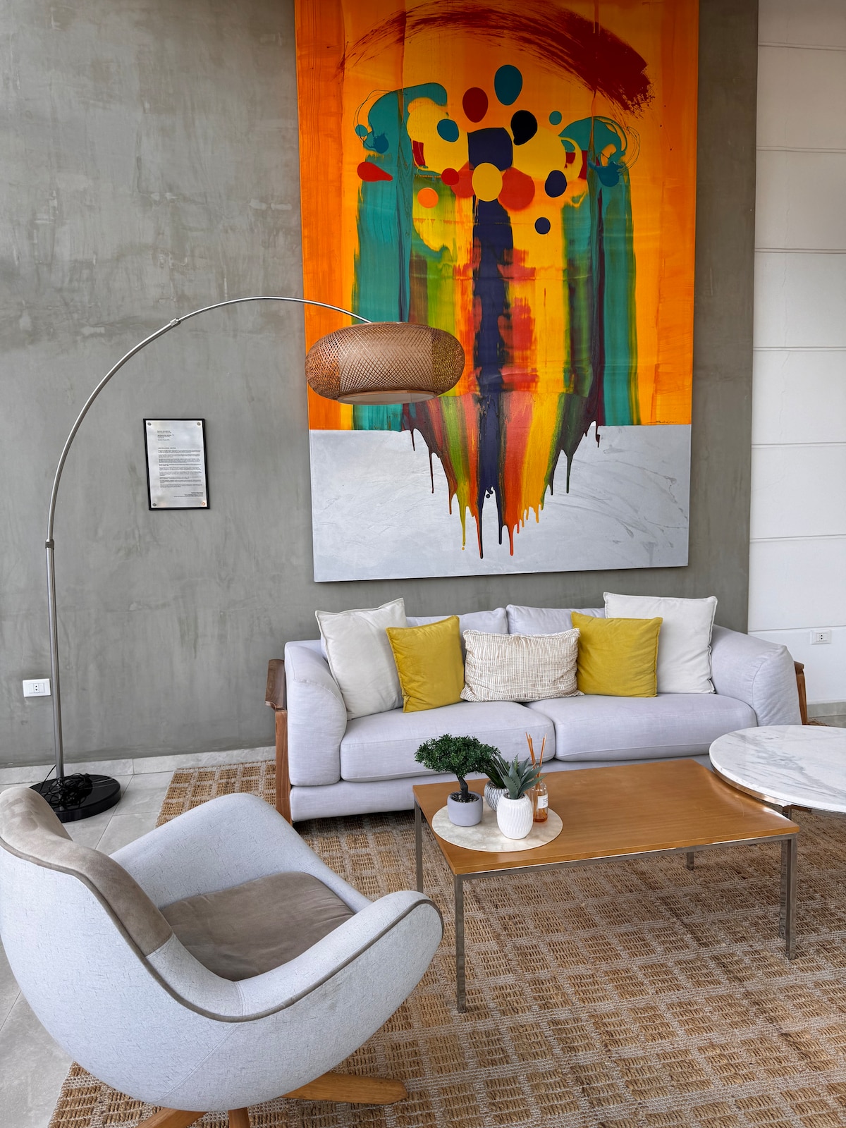 A modern living area features a light grey sofa adorned with yellow and white cushions, complemented by a wooden coffee table. A large, vibrant abstract painting hangs on the wall, and a stylish floor lamp provides ambient lighting. A cozy armchair enhances the inviting space.