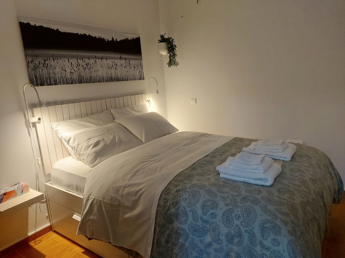 A spacious bed is presented with crisp white linens and a decorative bedspread featuring a subtle pattern. Two bedside lamps provide soft lighting, while a black-and-white landscape photograph adorns the wall above. Fresh towels are neatly arranged at the foot of the bed.