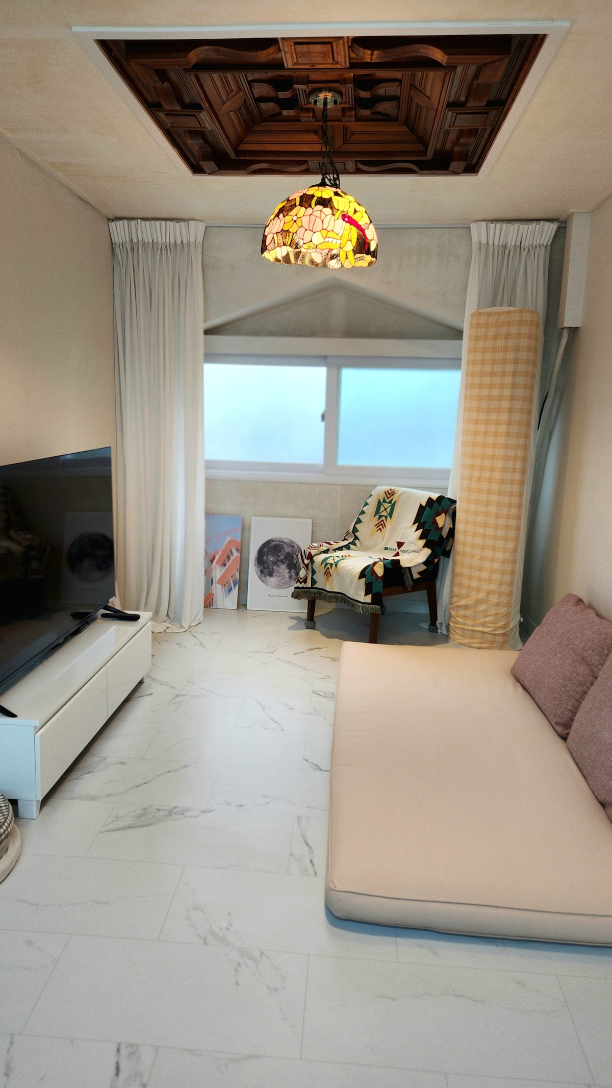 A cozy living area features a light-colored sofa and a wooden ceiling with an ornate design. A colorful stained glass pendant light hangs above, illuminating the space. A large window allows natural light to enter, complemented by soft drapes. Artwork is displayed on the floor.