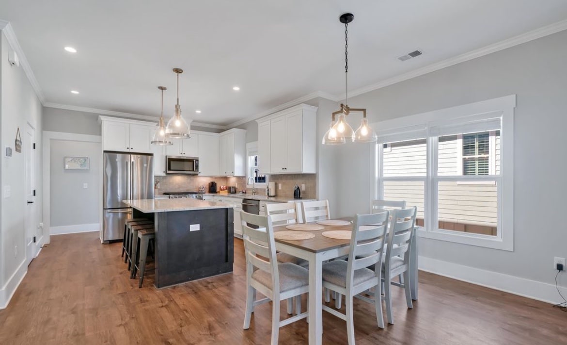 The modern kitchen area features white cabinetry and stainless steel appliances. An island with seating for four is complemented by a spacious dining table that seats six, all set against warm-toned hardwood floors and natural light illuminating the open layout.