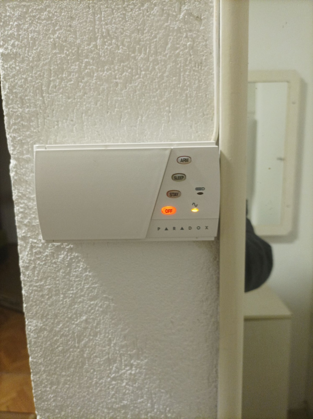 A wall-mounted control panel is positioned on a white textured surface. The panel includes several buttons and a small display, with a red indicator light illuminated, signaling its active status. A nearby mirror reflects part of the room, enhancing the image's context.