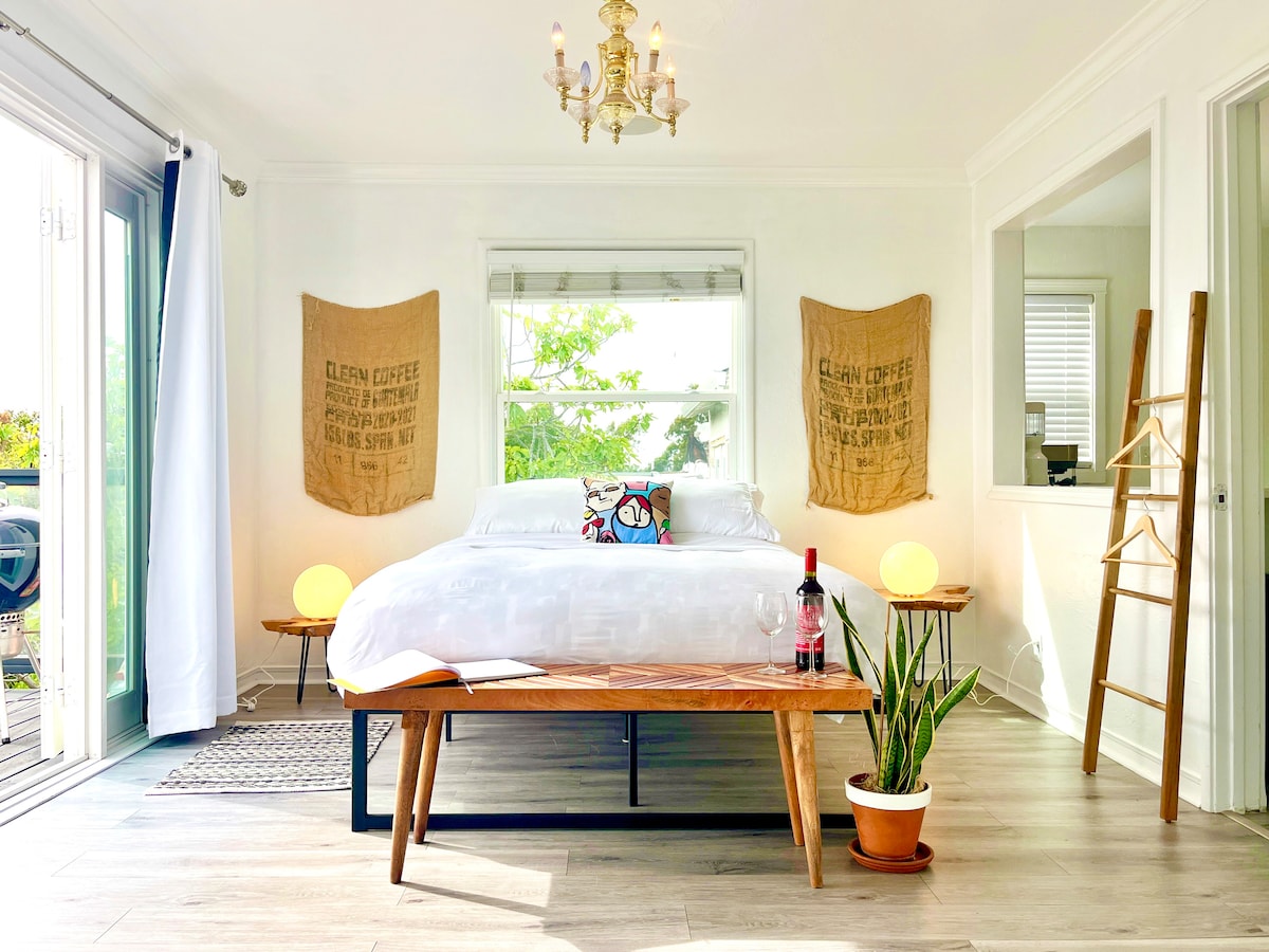 A bright bedroom features a spacious bed adorned with decorative pillows. Warm natural light enters through large glass doors, illuminating the minimalistic decor. A wooden bench sits at the foot of the bed, accompanied by a potted plant and soft lighting fixtures on either side.