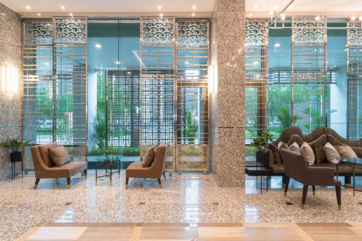An elegant lobby area features modern decor with comfortable seating arranged around small tables. Large glass doors provide a view of the greenery outside, while decorative screens add a touch of privacy. The polished floors reflect the ambient lighting, creating a welcoming environment.