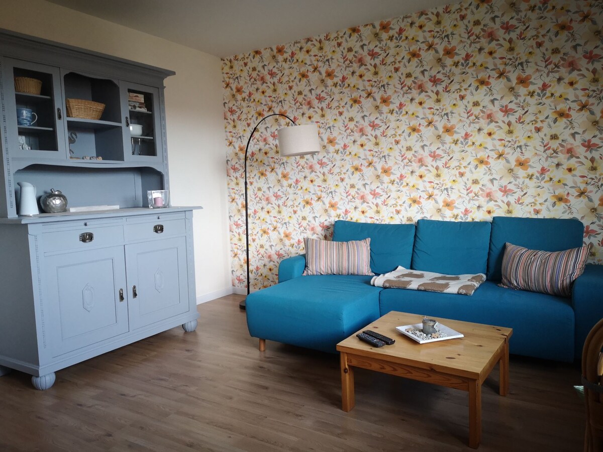 A living area features a light-colored wooden floor and a vibrant floral accent wall. A blue sectional sofa with soft cushions complements a wooden coffee table, which holds decorative items. A gray cabinet provides storage options, and a standing lamp adds ambient light.