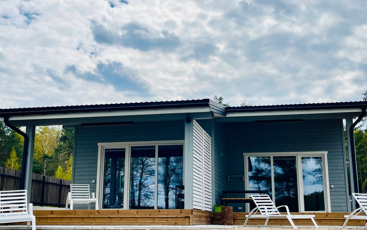 A modern wooden house features large glass doors that invite natural light into the space. A wooden deck extends in front, complemented by several white lounge chairs. The surrounding greenery and clouds add to the serene atmosphere.