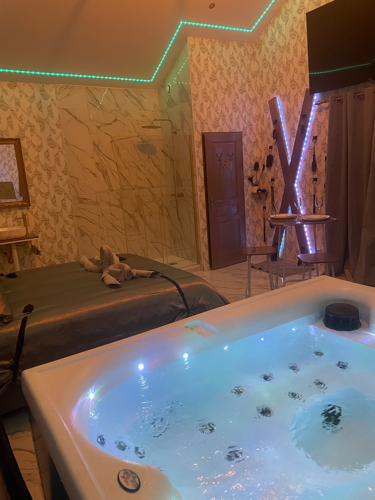 A luxurious suite features a spacious jacuzzi with illuminated water jets, accompanied by a well-made bed adorned with satin linens. Subtle lighting highlights decorative elements in the room, while a small bar area is visible in the background.