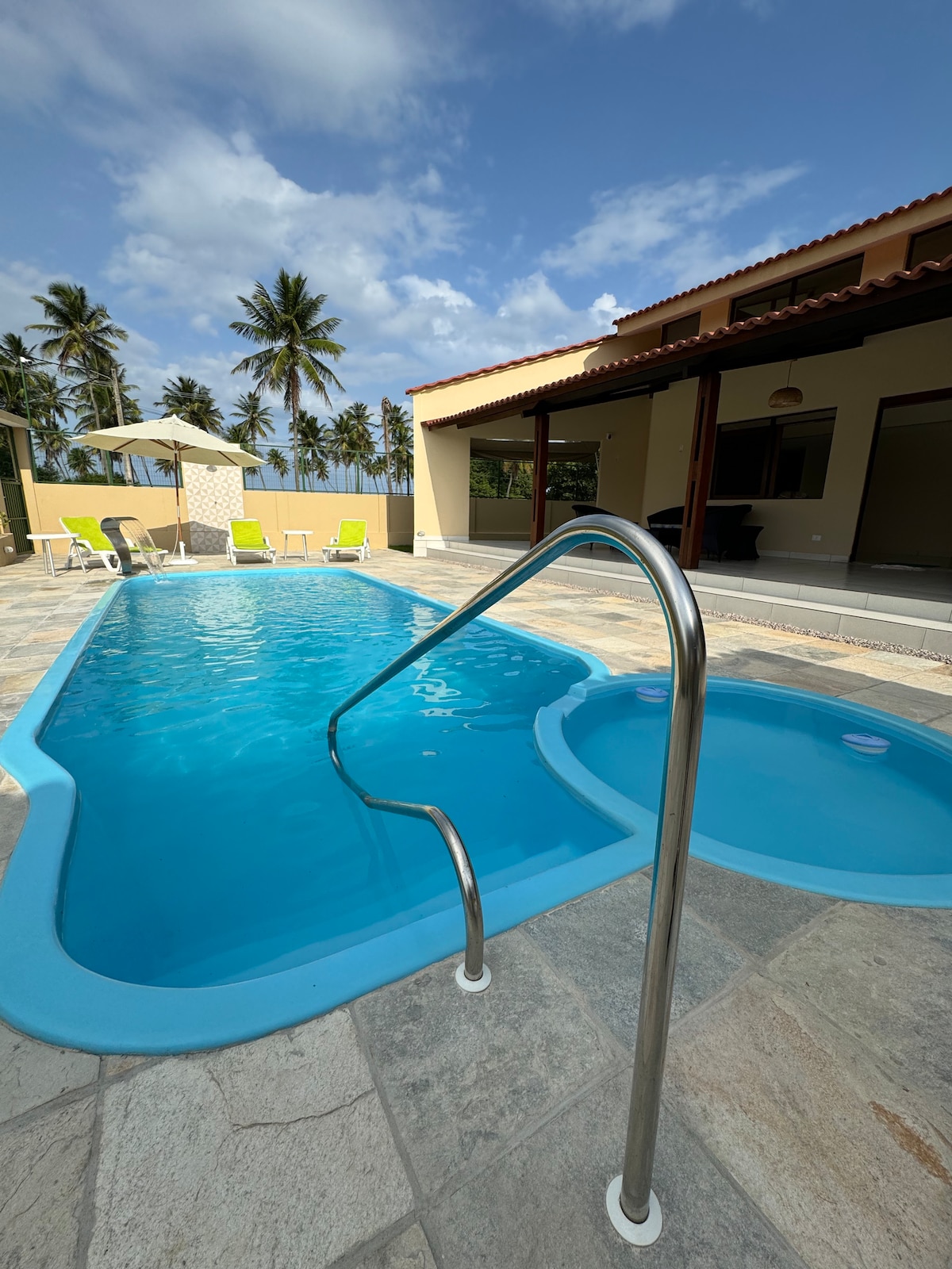 A swimming pool with a connecting hot tub is framed by palm trees under a partly cloudy sky. Lounge chairs in bright yellow are positioned around the pool area, while a shaded gazebo is visible in the background, offering a relaxing outdoor atmosphere.